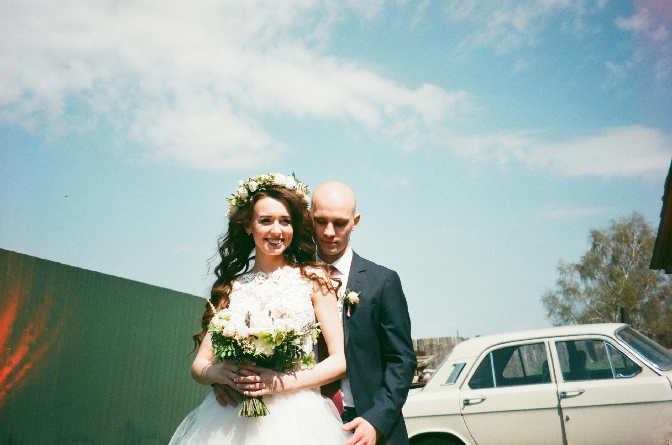 A joyful bride and groom embrace outdoors with a classic car in the background.