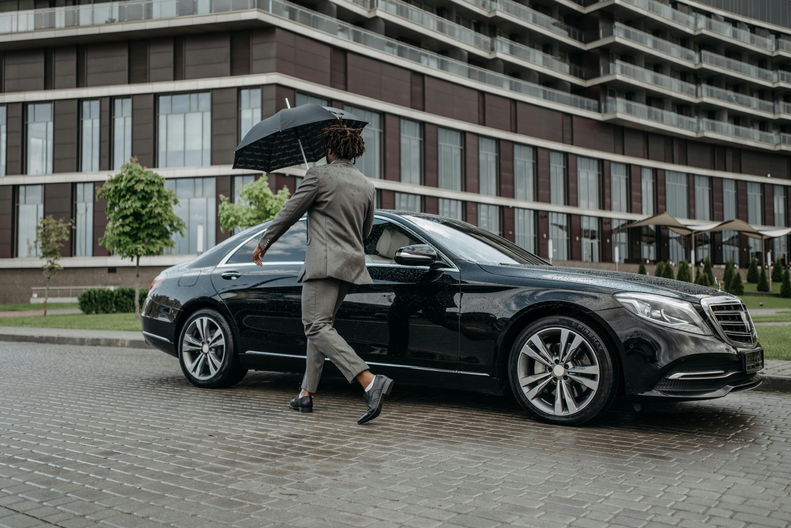 Man holding umbrella opens car door on rainy day in front of modern building.